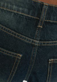 Mörkblå denimtyg med orange sömmar, som visar detaljerna för bakfickan och bältesslingan på jeans.