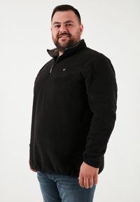 Fekete gyapjúpulóver félcipzárral, mellkasi kis logóval és textúrált anyaggal. Kék farmernadrág felett viselve.