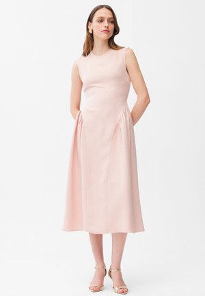 Donna in piedi che indossa un abito midi senza maniche di colore rosa chiaro con tasche, abbinato a sandali con tacco alto e cinturino alla caviglia dorati, su sfondo bianco.