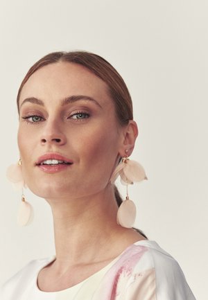 Heledanahaline naine, kellel on tagant kinni seotud juuksed, kannab suuri, pehmeid roosakate kroonlehekaedega kõrvarõngaid ja valget ülaosa roosade detailidega, vaadates kergelt ülespoole.