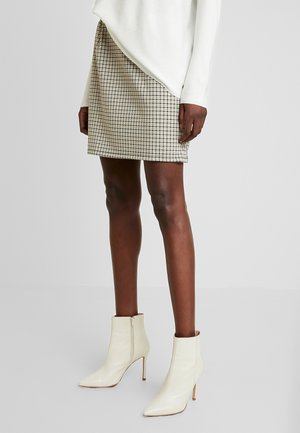 Persoon draagt een beige en zwarte geruite rok, een witte top met lange mouwen en witte puntige enkellaarsjes met hoge hakken, staand op een grijze vloer.