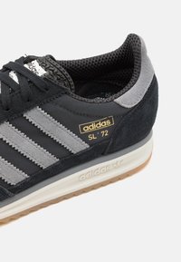 Svart och grå sportig sko i mocka och textil, med tre karakteristiska ränder och en guldfärgad logodetalj. Vit mellansula med gummisula.