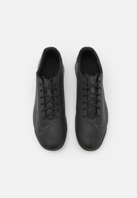 Zapatillas deportivas negras con un upper texturizado, que presenta un patrón en zigzag, acentos suaves y cordones negros. La suela es mínima y plana.