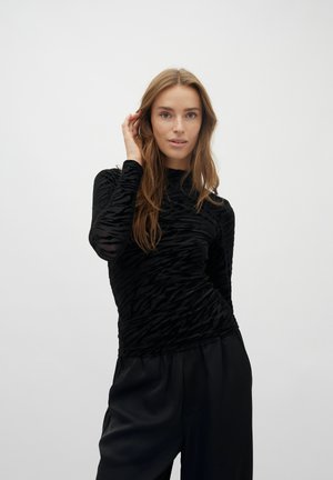 Mlada ženska z dolgimi lasmi, oblečena v črno majico z dolgimi rokavi in vzorcem ter sproščene črne hlače, stoji pred preprostim svetlim ozadjem.