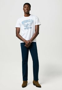 Camiseta de algodón blanca con gráfico azul "WRANGLER 1947", combinada con pantalones de mezclilla azul oscuro y botas de ante marrón hasta el tobillo.
