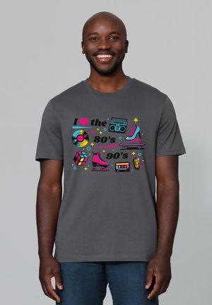 Lächelnder Mann mit grauem Hemd, das bunte Symbole der 80er und 90er zeigt: Schallplatte, Boombox, Rollschuh, Skateboard, Kassette, Mikrofon und Zauberwürfel.