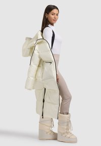 Puffet, off-white lang frakke med hætte og sort lynlås, båret over en hvid top og beige leggings. Beige vinterstøvler med snørebånd.