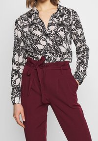 Schwarz-weiß gemusterte Bluse mit Kragen, die florale Designs aufweist. Kombiniert mit burgunderfarbenen Hochbundhosen mit einem gebundenen Bund.