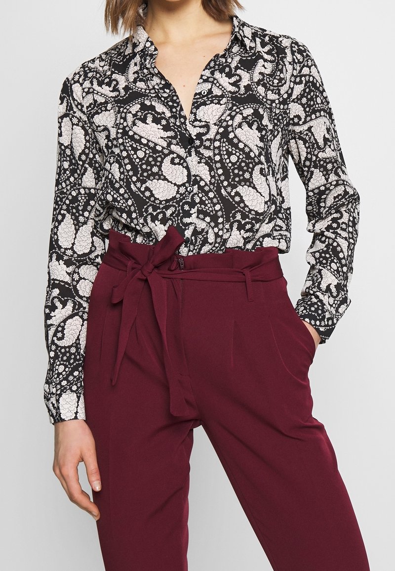 Schwarz-weiß gemusterte Bluse mit Kragen, die florale Designs aufweist. Kombiniert mit burgunderfarbenen Hochbundhosen mit einem gebundenen Bund.