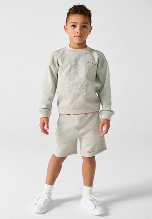 Boy standing wearing a light gray textured sweatshirt and matching shorts with white socks and white sneakers on a plain background.
