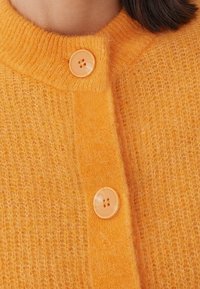 Orange, fluffig cardigan med ribbat halsband och två runda, ljusorange knappar framtill. Mjuk textur med ett varmt utseende.