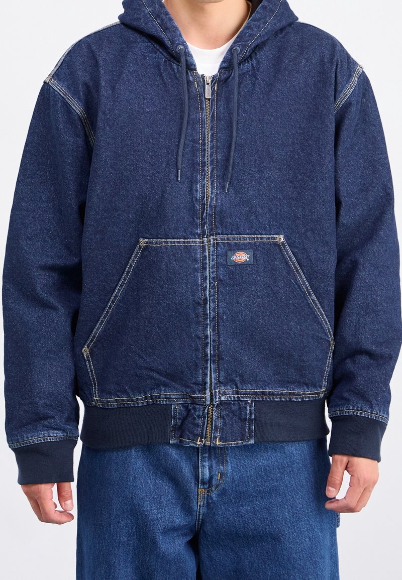 Veste à capuche en denim bleu avec fermeture éclair frontale, grandes poches avant, poignets et ourlet côtelés, avec des coutures contrastantes.