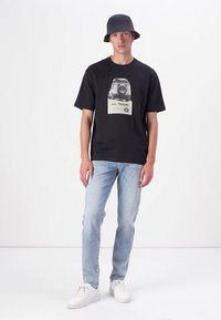 Camiseta negra de manga corta con un gráfico de un vehículo, combinada con jeans de mezclilla azul claro y zapatillas blancas. El modelo lleva un sombrero tipo cubo oscuro.