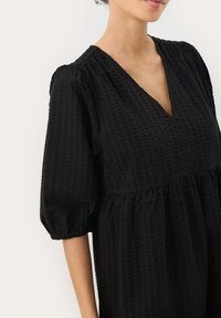 Zwarte gekeerde jurk met een V-hals, korte pofmouwen en een geraffineerde taille. De stof heeft een subtiel ruitpatroon.