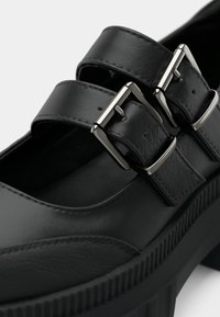 Chaussure en cuir noir avec deux brides à boucle, bout arrondi et semelle plateforme texturée avec détails rainurés.