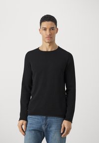 Tricou negru cu mânecă lungă, realizat dintr-un material moale, cu un guler rotund și croială ajustată. Asortat cu blugi de culoare albastru deschis.