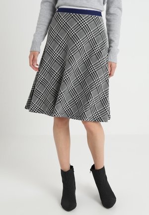 Kvinde iført sort-hvid ternet knælang nederdel, grå sweater og sorte ankelstøvler, står på hvid baggrund.
