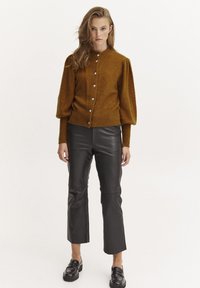 Cardigan lavorato a maglia color senape con maniche a sbuffo, bottoni argentati sul davanti, indossato sopra pantaloni di pelle nera e mocassini neri.