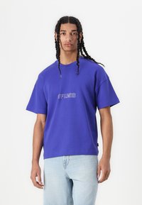 T-shirt oversized violet en coton, avec un col rond et des manches courtes. Le texte graphique à l'avant indique "HORS LIMITES" en blanc.