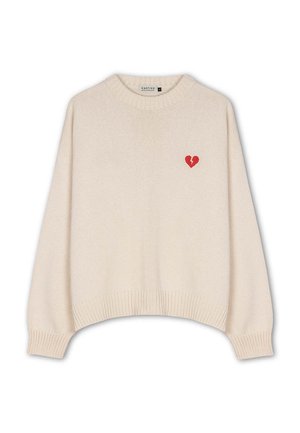 Bežový pletený sveter s rebrovanými manžetami a lemom, s malou červenou zlomenou srdcovou emblematickou nášivkou na hornej ľavej strane hrude.