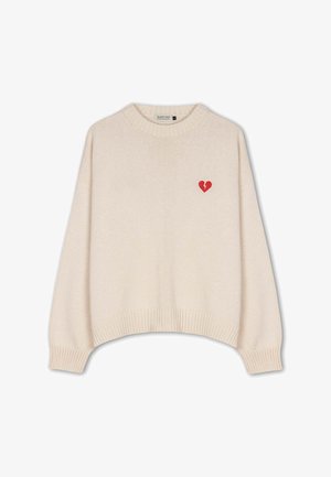 Bežani pleteni pulover s rebrastim rukavima i rubom, sa malim crvenim simbolom slomljenog srca na gornjem lijevom dijelu prsiju.