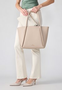 Beige tygväska i pebblat läder, med två handtag. En liten vit påse är delvis synlig inuti. Modellen bär ljusa krämfärgade byxor och skor.