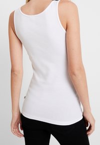 Débardeur en coton blanc avec un col rond, coupe ajustée et fines bretelles. Texture lisse et étiquette de marque discrète sur le côté.