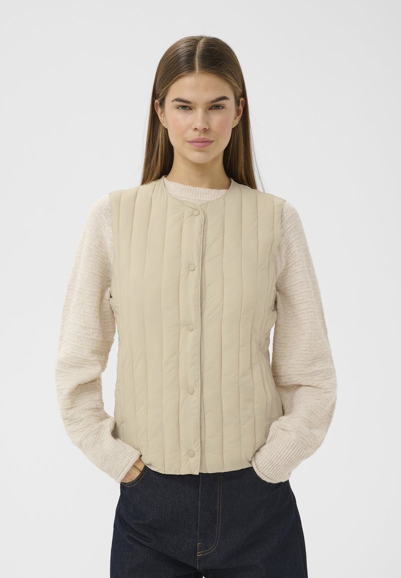 Jeune femme portant un gilet matelassé beige sur un pull crème texturé et un jean bleu foncé, debout les mains dans les poches devant un fond blanc.