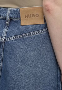 Patch en cuir marron avec "HUGO" sur un jean en denim bleu porté par une personne avec un haut beige et un bras visible.