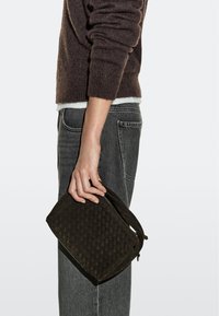 Personne portant un pull marron foncé et un jean gris tenant une pochette tissée marron à côté d'eux sur un fond uni.