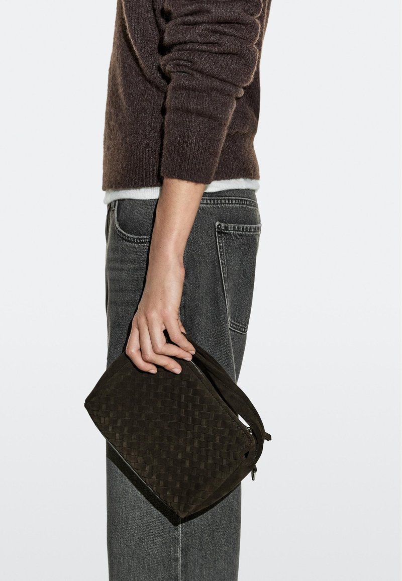 Personne portant un pull marron foncé et un jean gris tenant une pochette tissée marron à côté d'eux sur un fond uni.