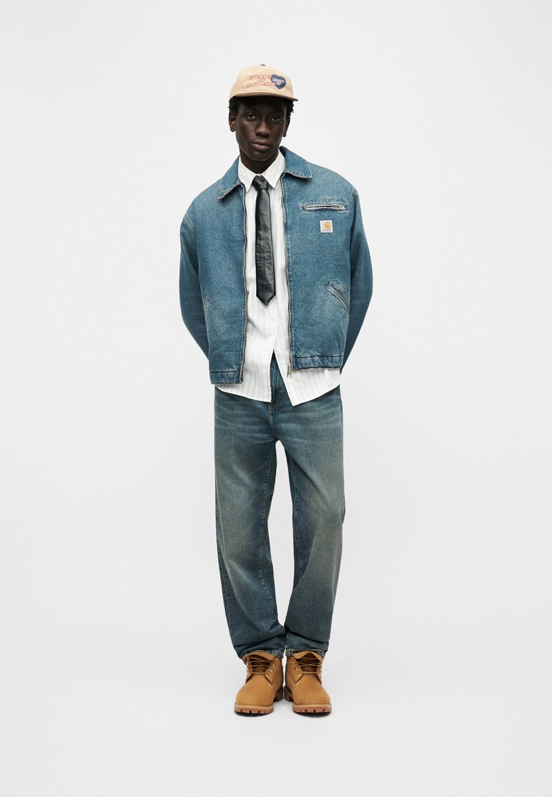 Homme portant une casquette beige, une veste en jean bleue, une chemise blanche avec une cravate noire, un jean bleu et des bottes de travail marron clair sur fond blanc.