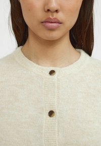 Crèmekleurige gebreide cardigan met een ronde halslijn, voorzien van een textuur ribbelrand en een ronde bruine knoopsluiting.