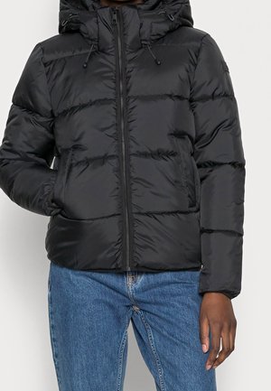 Osoba ubrana w czarną pikowaną kurtkę z kapturem, zapinaną na zamek z przodu i kieszeniami, w zestawieniu z niebieskimi dżinsami na neutralnym tle.