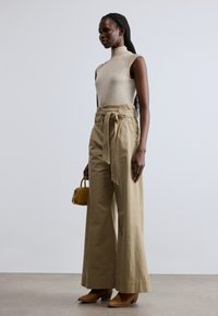 Top bej fără mâneci, cu guler înalt, pantaloni kaki largi talie înaltă cu curea legată, cizme maro cu toc și geantă mică galbenă.