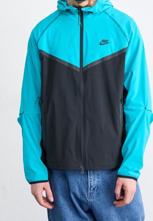 Personne portant une veste à capuche Nike turquoise et noire avec une fermeture éclair à l'avant, assortie à un jean bleu, faisant face à l'avant.