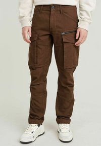 Bruine cargobroek met meerdere zakken, een ritssluitingszak aan de zijkant, stevige stof, taps toelopende benen en een knoopsluiting. Verstelbare tailleband.