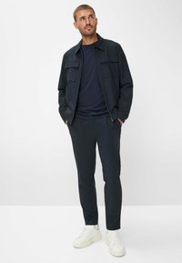 Marineblauwe jas met borstzakken over een bijpassend t-shirt, gecombineerd met strakke marineblauwe broek en witte sneakers. Soepele stoftextuur, moderne pasvorm.