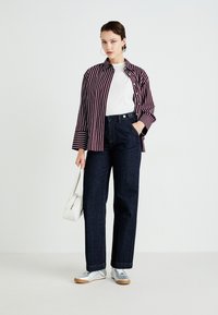 Donna con jeans a gamba larga di un blu scuro, t-shirt bianca, camicia a righe bordeaux e bianca, sneakers argento, che tiene una borsa bianca, in piedi contro uno sfondo neutro.