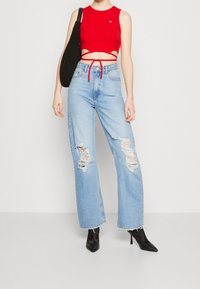 Top de tirantes rojo cortado con detalles recortados, combinado con vaqueros cuyas perneras son anchas y rasgadas en un tono azul claro. Botas de tacón negras con punta afilada y un bolso negro.
