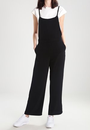 Schwarzer Overall mit dünnen Trägern, weitem Bein Design, Taschen, kombiniert mit einem weißen T-Shirt und weißen Sneakern, vor einem neutralen Hintergrund.