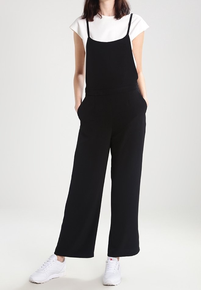 Combinaison noire à fines bretelles, design à jambes larges, poches, associée à un t-shirt blanc et des sneakers blanches, sur un fond neutre.