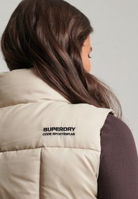 Krämfärgad vadderad väst med quiltad design, med en svart "SUPERDRY CODE SPORTSWEAR"-logga på baksidan.