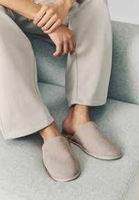 Beige Strickschlappen mit strukturiertem Obermaterial und weicher Sohle, kombiniert mit hellgrauen, lockeren Hosen, auf einer grauen Oberfläche sitzend.