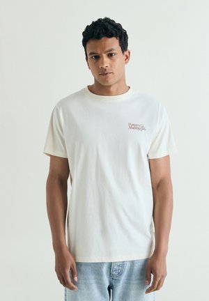 Hombre con cabello corto y rizado que lleva una camiseta blanca con el texto "Pottery Making" y vaqueros azul claro, de pie frente a un fondo liso.