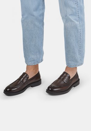 Mocasines de cuero marrón con un acabado suave, punta redonda y suela baja de goma. Detallados con una correa horizontal en el empeine.
