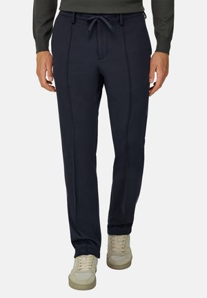 Homme portant un pantalon slim bleu marine avec une taille à cordon et des baskets beige, debout devant un fond uni.