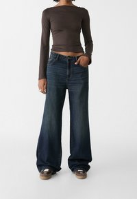 Hnědý dlouhý rukáv top v kombinaci se širokými tmavě modrými džíny. Džíny mají vysoký pas a jemné vyblednutí. Obuv zahrnuje hnědé tenisky.