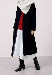Lange zwarte jas, oversized met een dubbelzijdig knoopontwerp. Gelaagd over een rode trui en een witte rok. Gecombineerd met zwarte laarzen tot over de knieën.