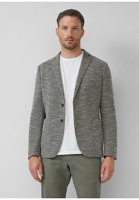 Herenblazer in grijs, met een gestructureerde stof, notchrevers, twee knopen en twee voorzakken. Draag het over een witte shirt en groene broek.